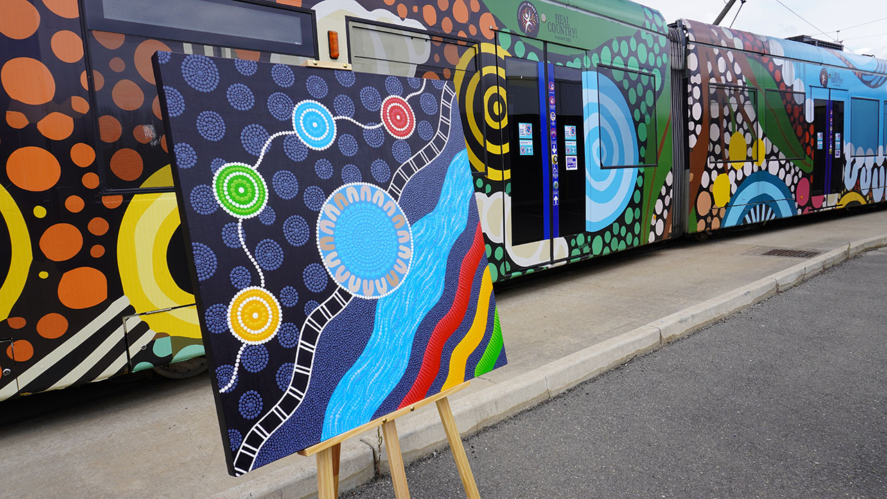 A brightly coloured Aboriginalpainting on an eisel in front a tram wrapped in an Indigenous design.