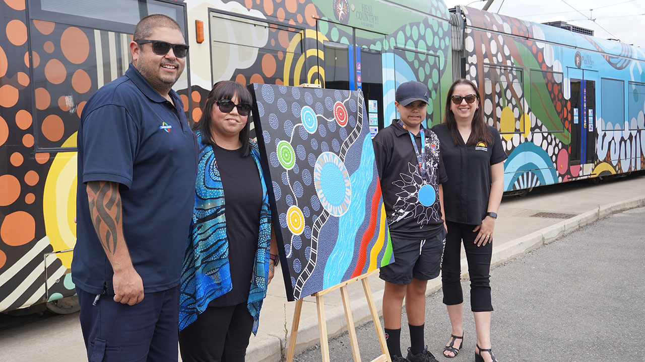 Andrew Sumner and artist Milly Sumner stand to the left of a painting on an eisel with their son and Rebecca Wessels standing to the right. A tram wrapped in Indigenous artwork is behind them.
