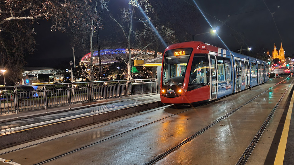 A red and blue Citadis tram sits at the Festival Plaza tram platform under a night sky with a birightly lit Adelaide Oval and St Peter's Cathedral behind it.