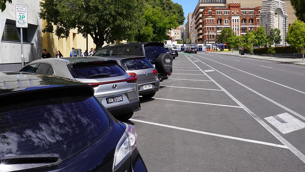 A row of cars parallel-parked on the street with a tra crossing an intersection in the background.