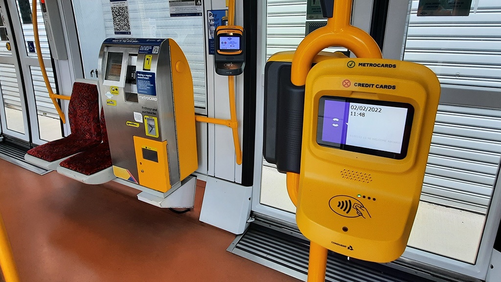 A yellow smart validatorin the foreground, with a grey validator in the background next to an on-board ticket vending machine.